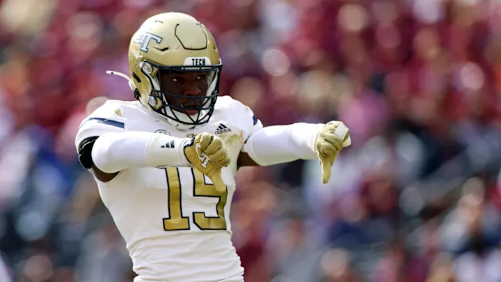 Georgia Tech football player wearing number 15 in white away uniform and gold helmet making downward pointing gestures with both hands during a game, with blurred crowd in stadium background
